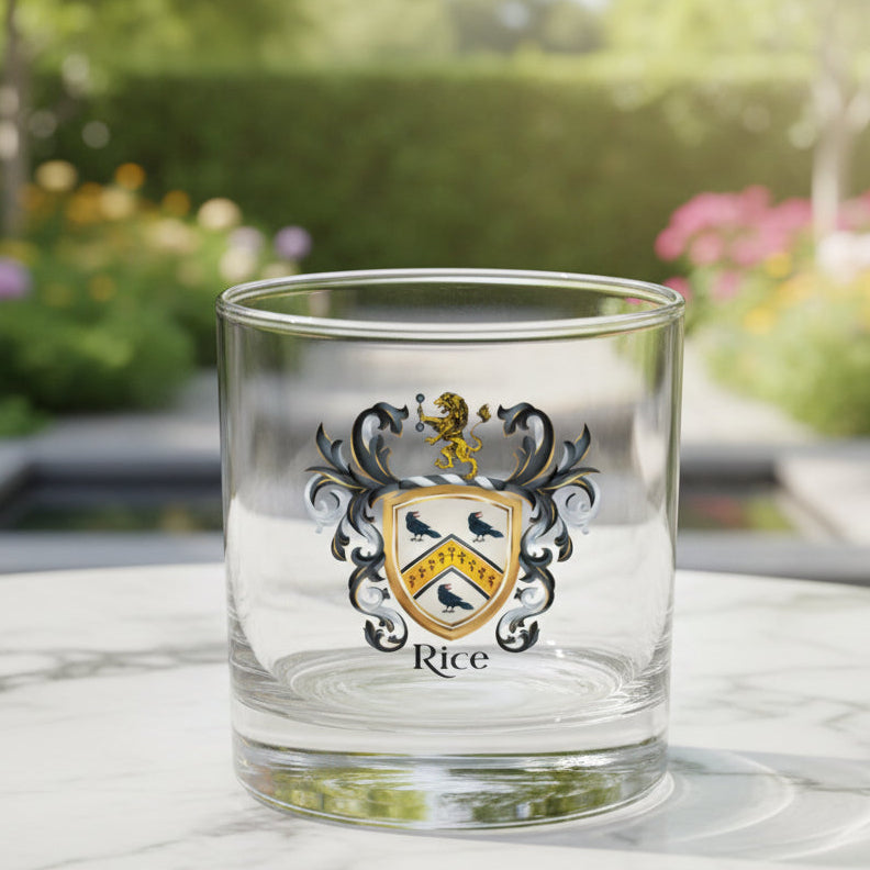 A clear glass rocks tumbler featuring a custom printed family crest with the name 'Rice' below it. The crest includes a gold lion, black and white mantling, and a shield with black birds on a gold background, placed on a white marble table top with a contemporary garden in the background on a sunny day.