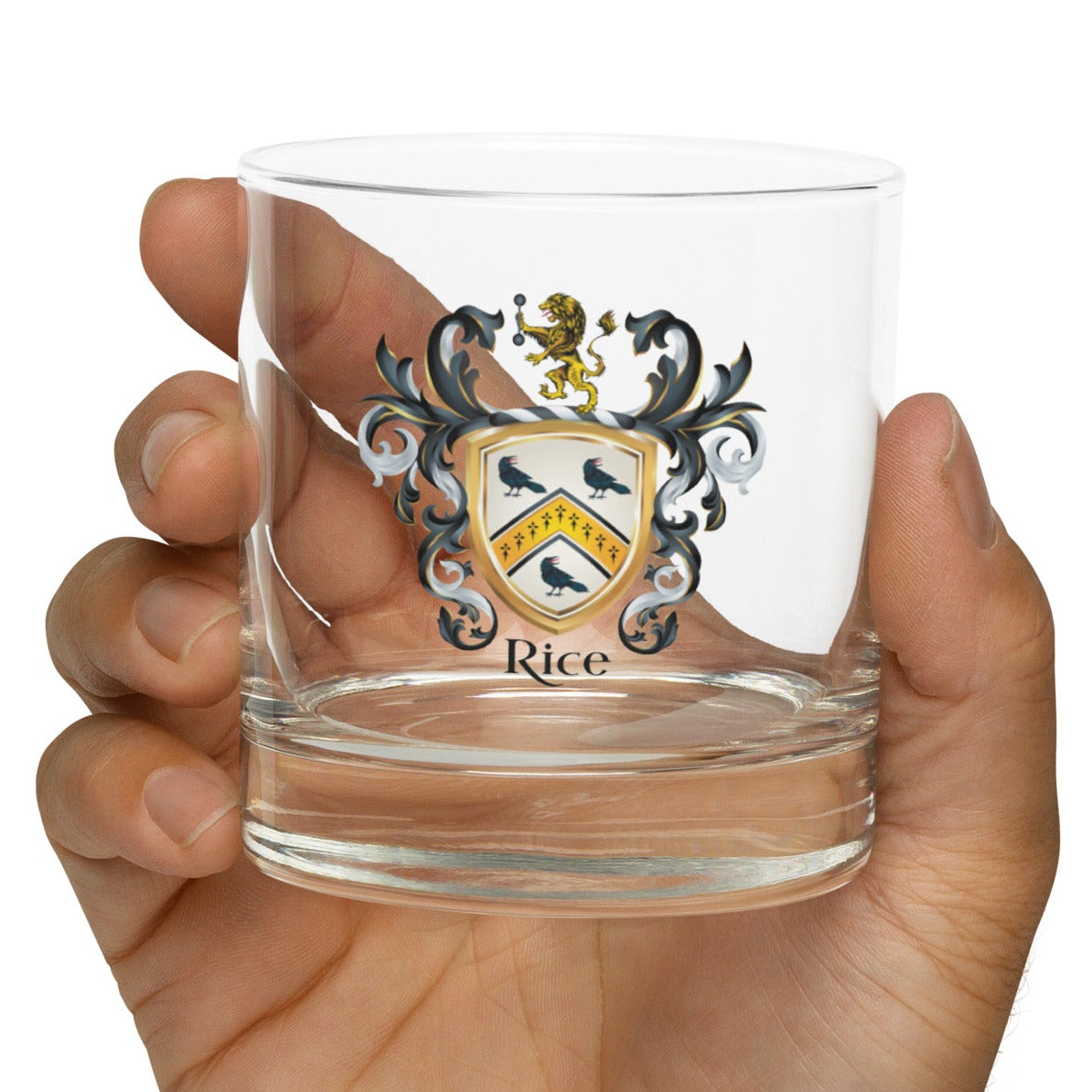 A hand holding a clear glass rocks tumbler featuring a custom family crest with the name 'Rice' printed below it, set against a plain white background.