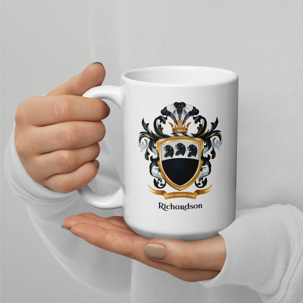 A person in a white long-sleeve top holds a white ceramic mug with a custom family crest design. The crest features a black and gold shield with three black lion heads, a black and white helmet, and the name 'Richardson' printed below in black.