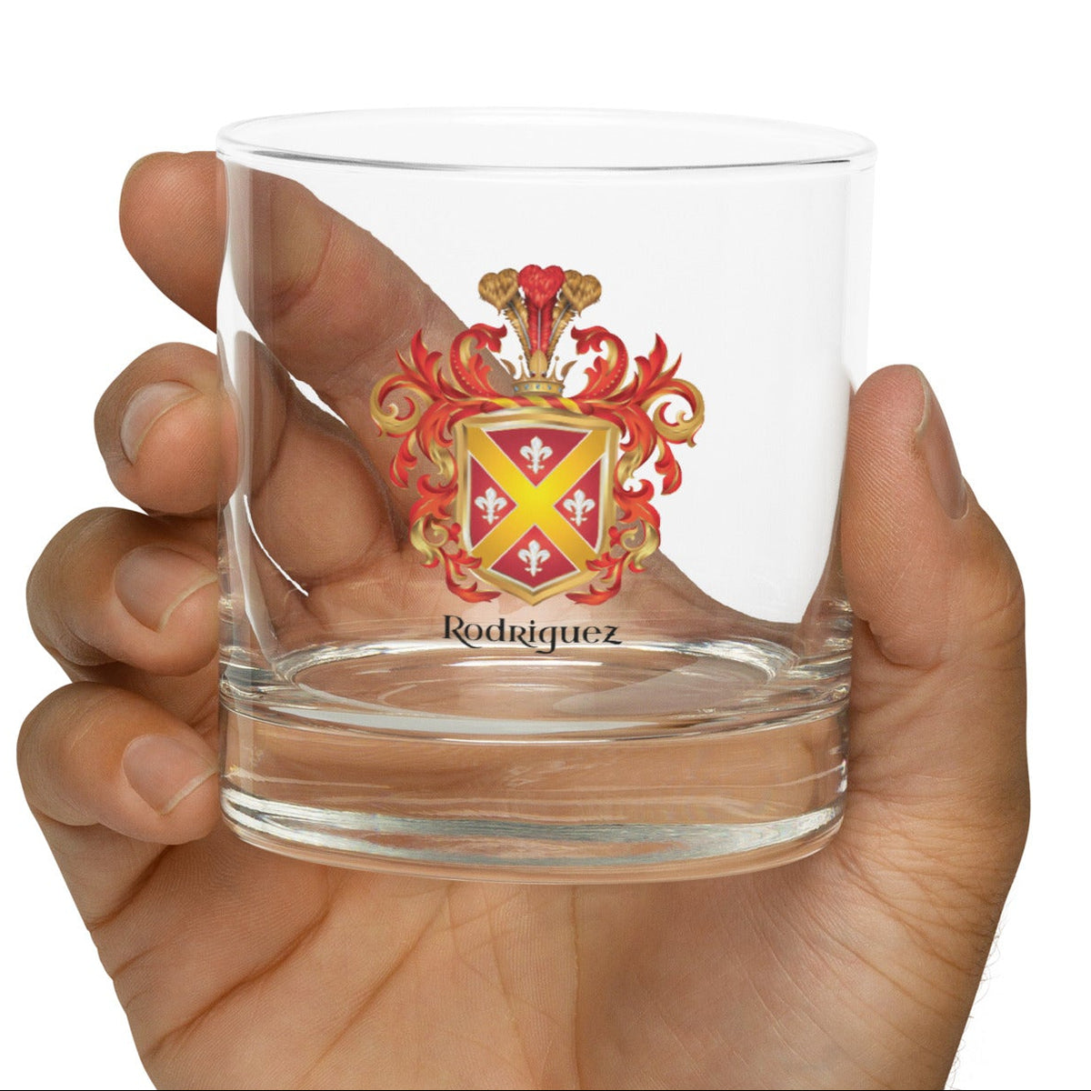 A hand holding a clear glass rocks tumbler featuring a printed red and gold Rodriguez coat of arms crest with the name 'Rodriguez' below it, set against a plain white background.
