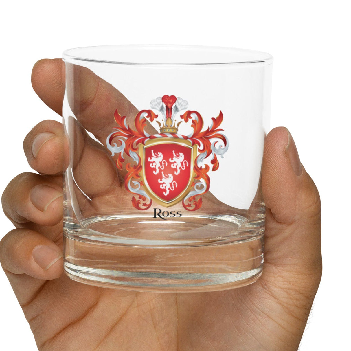 Hand holding a clear rocks glass with a Ross family crest with 3 white lions on a red shield on a white background