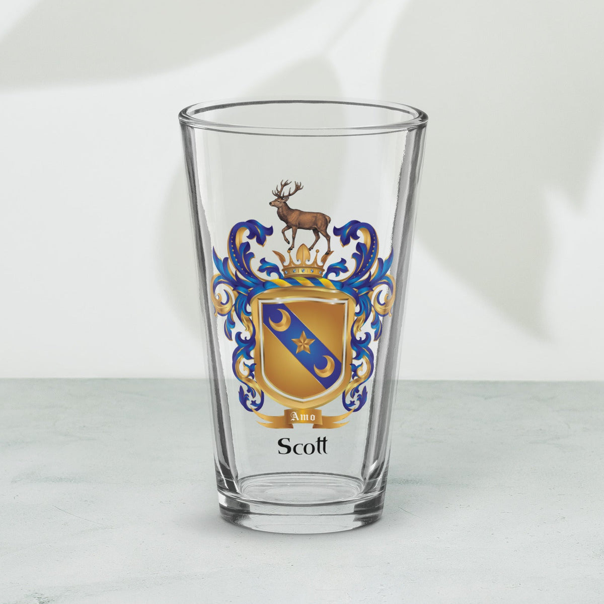 A clear pint glass with a custom family crest printed on the front, featuring a gold and blue shield with a stag and the name 'Scott' printed below. The glass is set on a light gray marble surface against a white wall with soft leaf shadows.