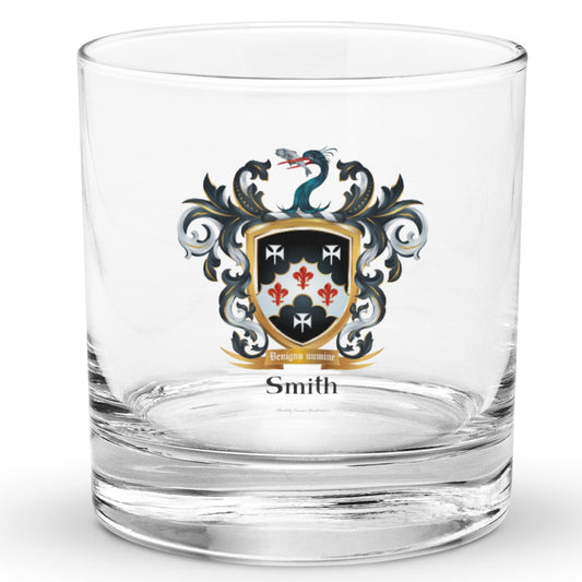 A classic rocks glass on a white background with a Smith  Smith Family Crest on the front that features a black shield with three white stylised crosses and theree red fleur de lys and also has a heron's head holding a fish in its beak on the crest above the shield.