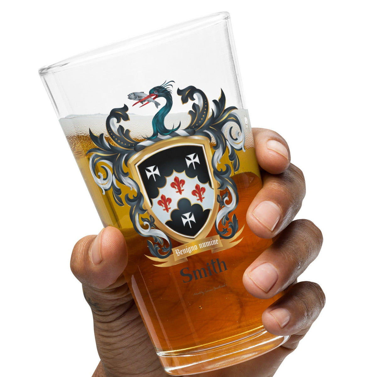 A hand holding a pint glass with the English Smith Family Crest on the front that features a black shield with three white stylised crosses and three red fleur-de-lys and also has a heron's head holding a fish in its beak on the crest above the shield.