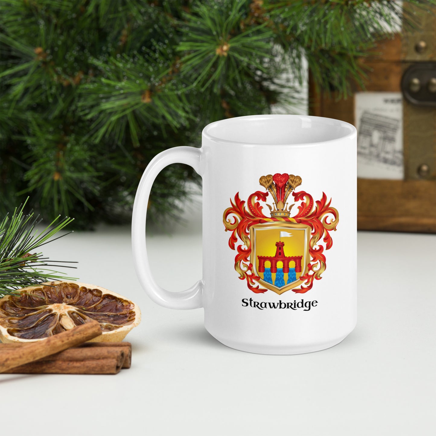 A white ceramic mug with a colorful Strawbridge Family Crest printed on the front, styled on a tabletop with pine branches, a dried orange slice, cinnamon sticks, and a rustic wooden box in the background.