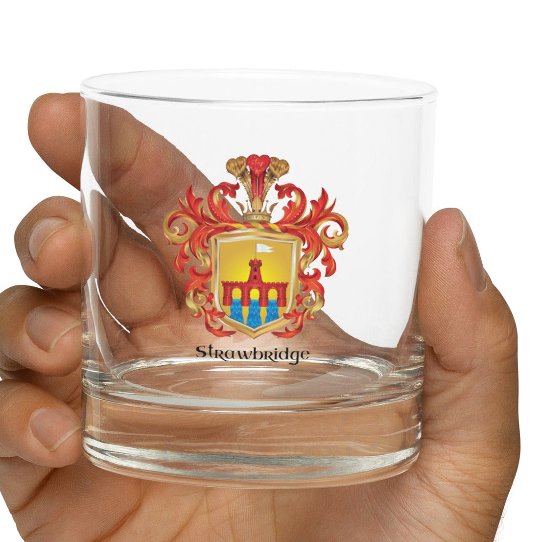 A hand holding a clear glass rocks tumbler featuring a colorful coat of arms with the text 'Strawbridge' printed below it, set against a plain white background.