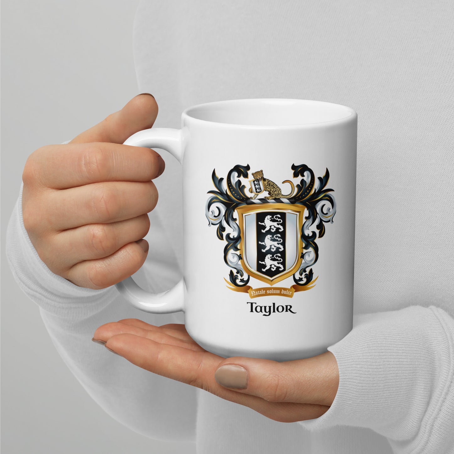Two hands holding a white mug with a Taylor family crest and the name 'Taylor' in black text below the shield that has 3 white lions on a black and white shield, the person is against a gray background