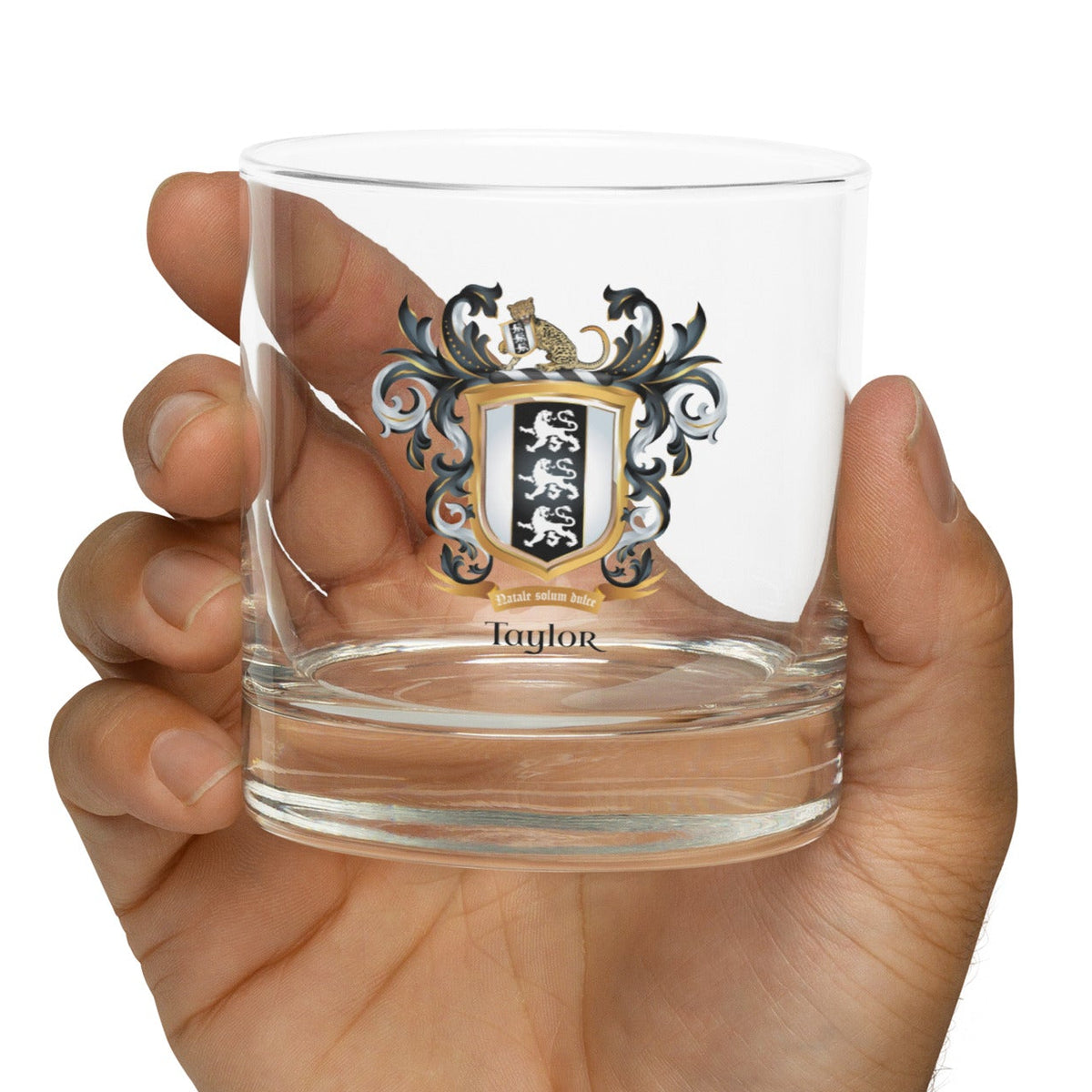 Hand holding a clear rocks glass with a aylor family crest and the name 'Taylor' in black text below the shield that has 3 white lions on a black and white shield on a white background
