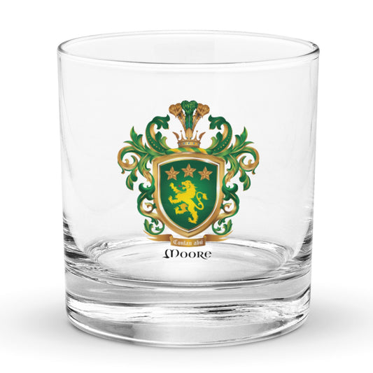 A clear rocks glass featuring The Moore Family Crest showing a green shield with a gold lion and three gold stars above the lion with the motto and Moore name beloe the shield and a crown above and green and gold mantling all around  on a white background