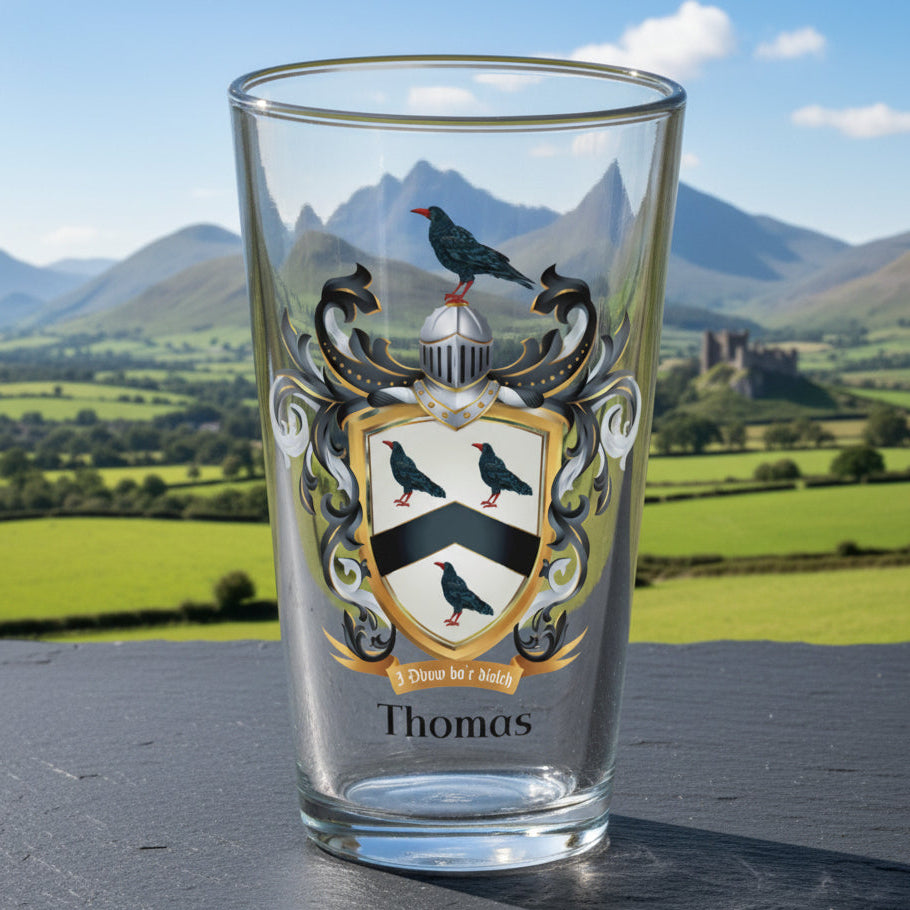 Thomas Family Crest beer glass on a black slate tabletop with a mountainous Welsh rural scene with a castle in the far distance on a sunny day.