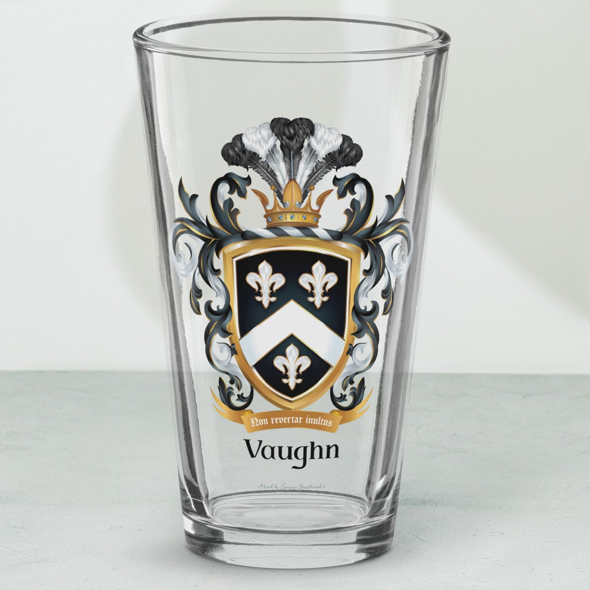 A clear pint glass with a custom printed Vaughn family crest on the front, set on a light gray marble surface with soft leaf shadows cast on a white wall behind it.