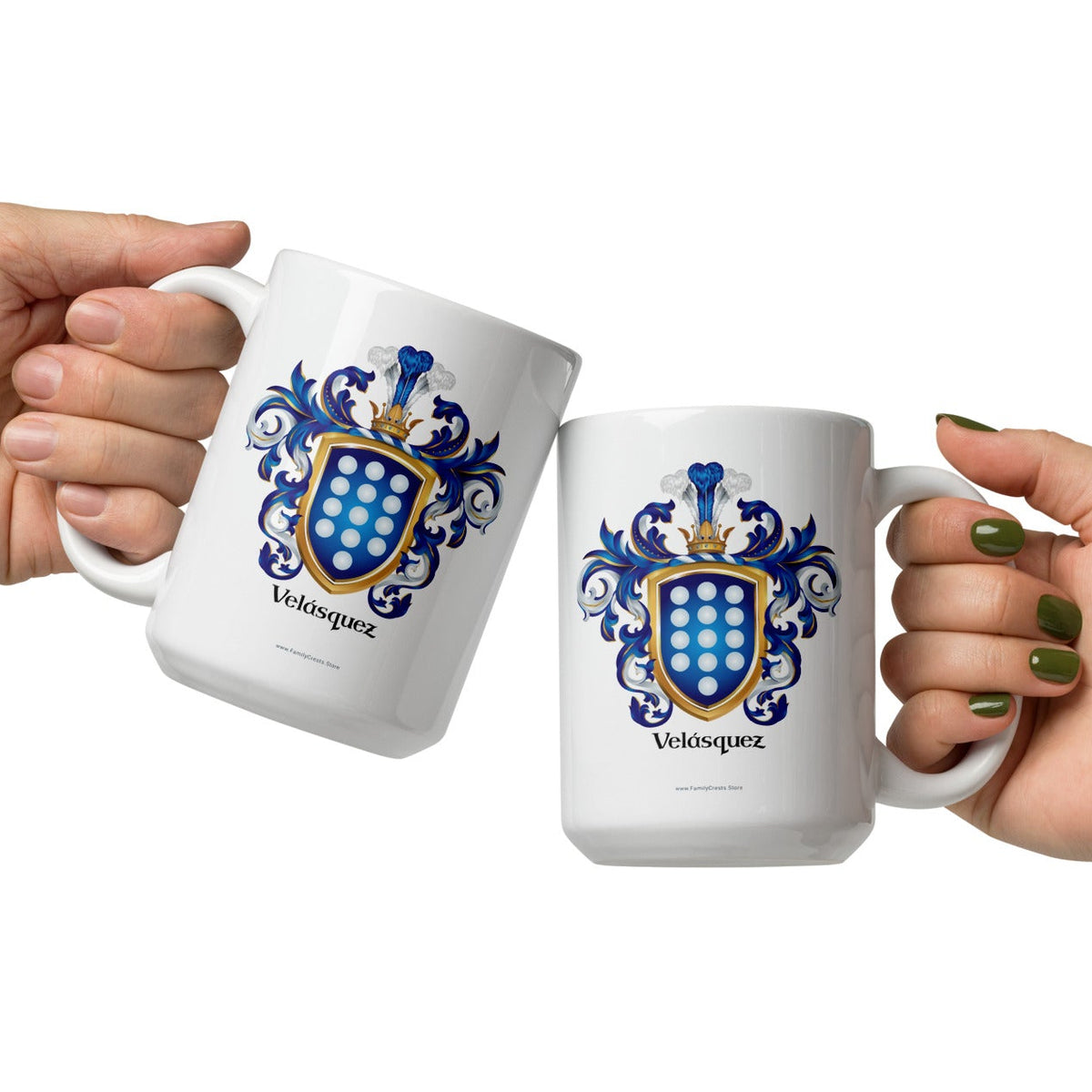 Two hands holding white ceramic mugs with a blue and gold Velasquez coat of arms design, set against a plain white background.