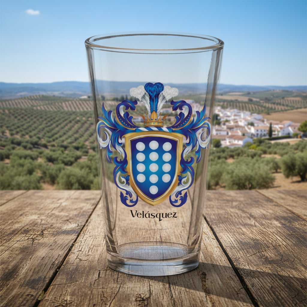A clear pint glass featuring a printed blue, gold, and white Velásquez family crest with the name 'Velásquez' printed below in black, shown on an old wooden table with a rural Spanish scene with olive trees in the far distance