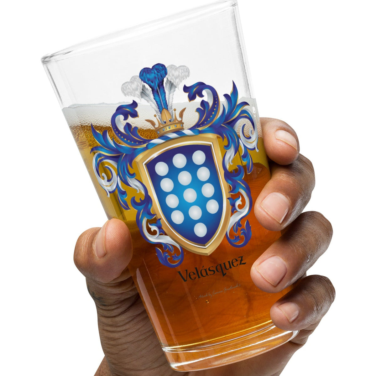 A hand holding a clear pint glass with a blue, white and gold Velasquez coat of arms design and amber beer inside, against a plain white background.