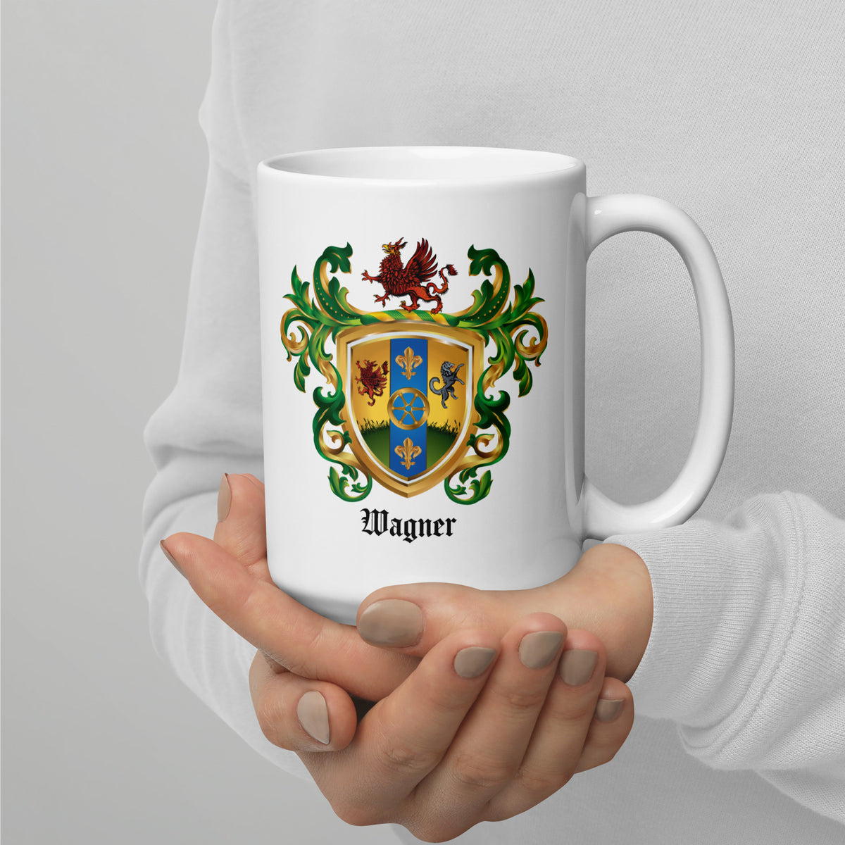 A person holding a white ceramic mug with a colorful family crest design and the name 'Wagner' printed below it, wearing a light gray long-sleeve top.