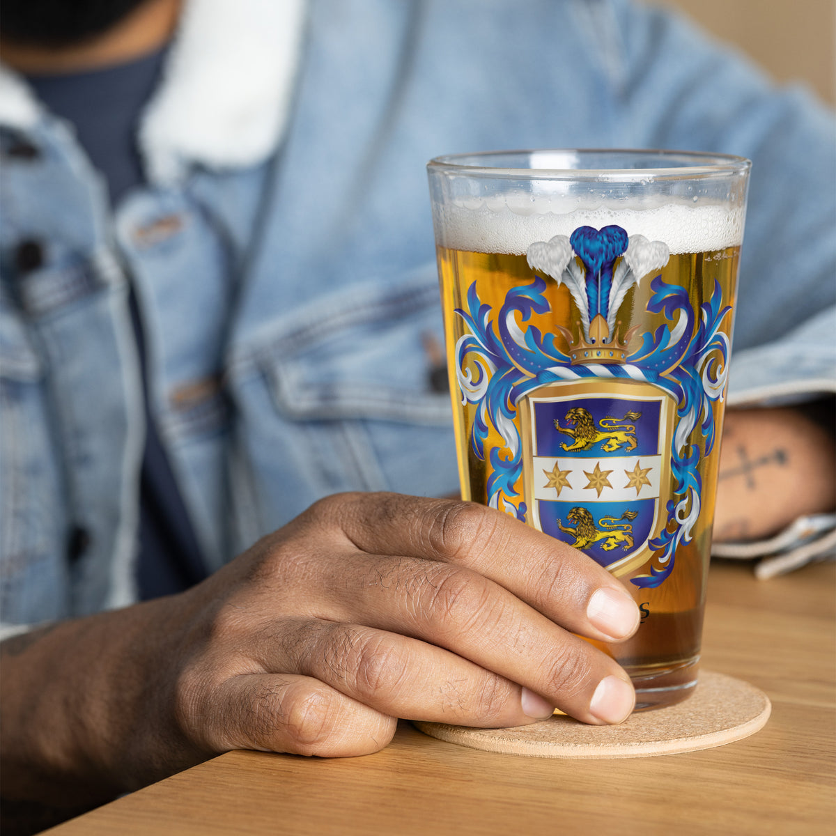 A hand holding a pint glass with a colorful heraldic crest design, filled with beer and foam, resting on a coaster on a wooden table.