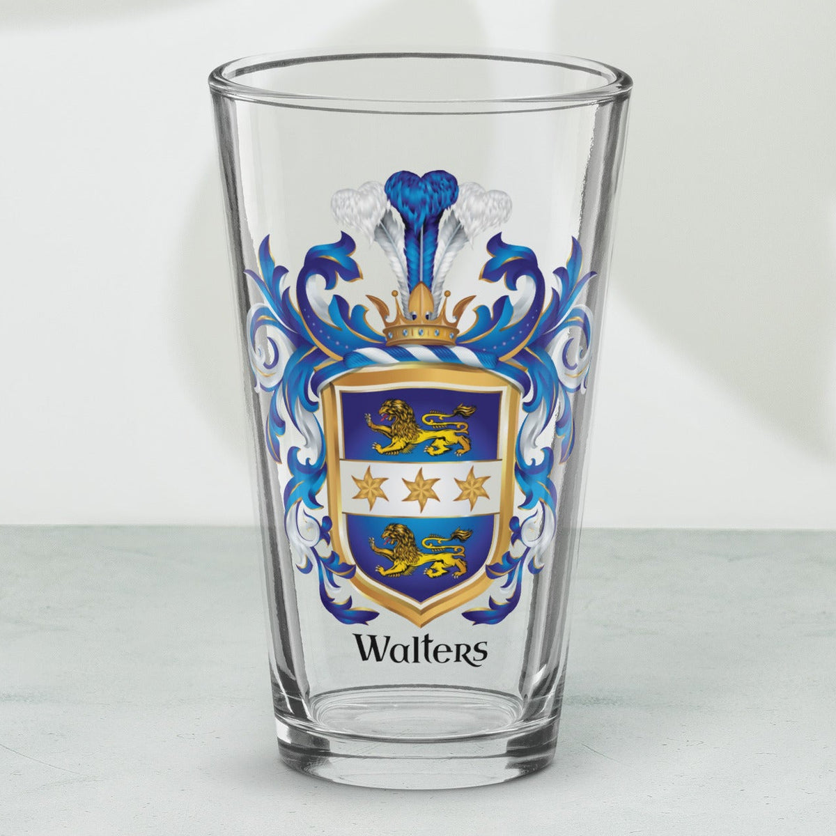 A clear pint glass with a custom printed family crest featuring blue, gold, and white details, and the name 'Walters' printed below, set on a light gray marble surface with soft leaf shadows.