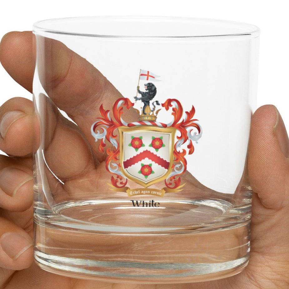A hand holding a clear glass rocks tumbler with a printed illustration of the White family crest and the name 'White' visible under the shield in black lettering, against a plain white background.