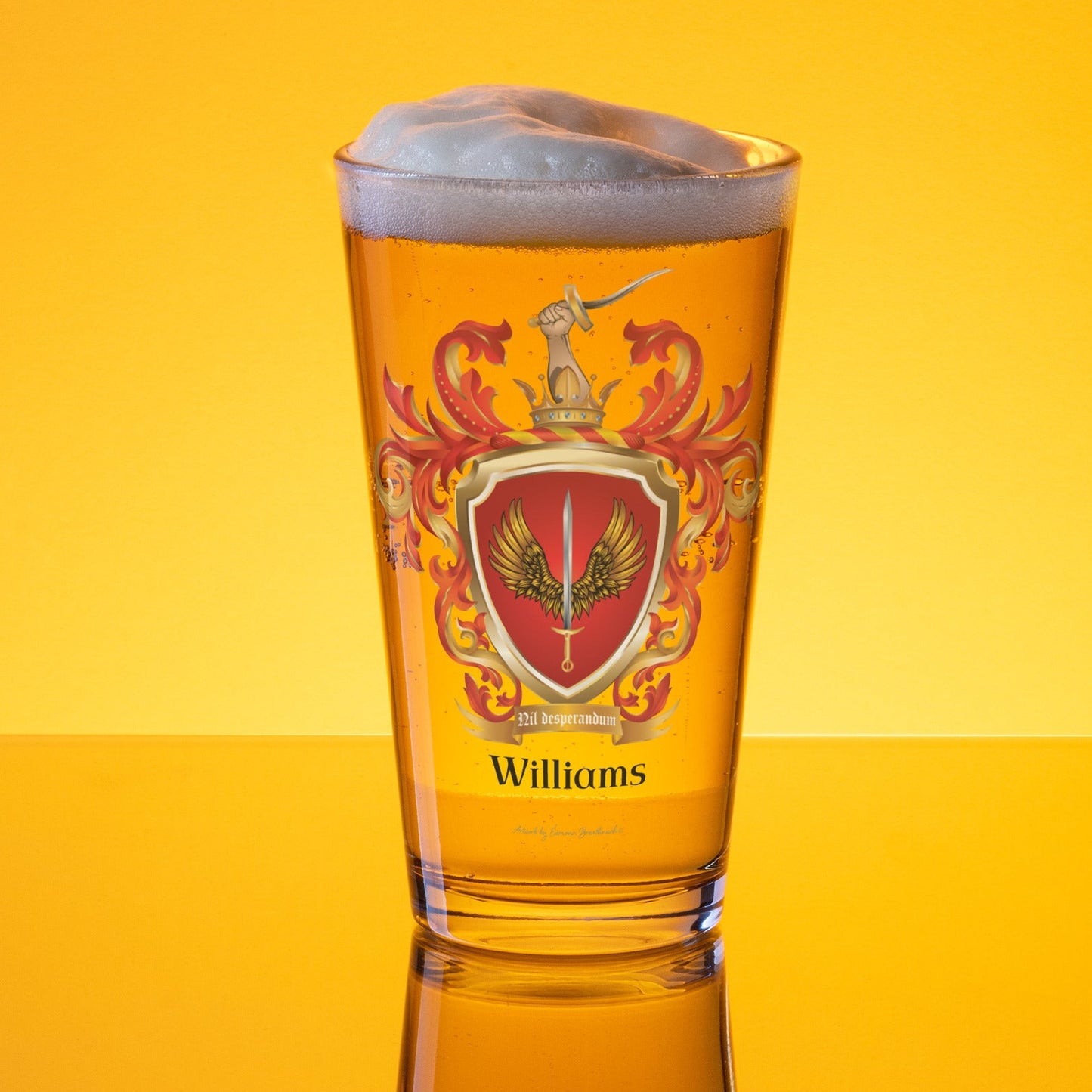 Williams Family Crest pint glass against a a golden lit background with the coat of arms on the glass showing a sword between two golden wings on a red shield and a hand with a sword coming out of a crown at the top on the crest 