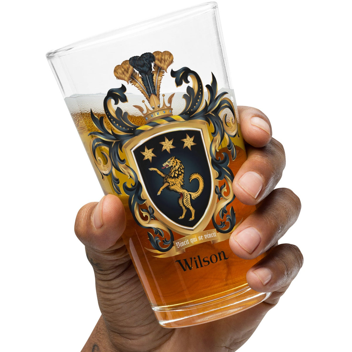 Hand holding a glass of beer with a AWilson Family Crest featuring a golden wolf with 3 stars on a black shield photographed on a white background.