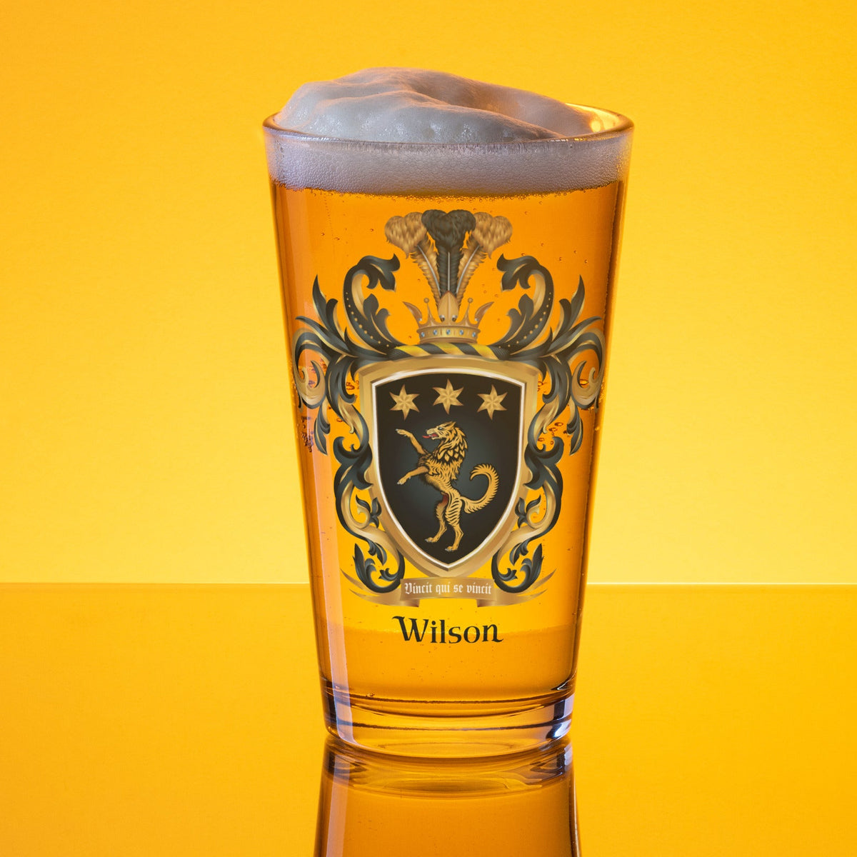 A clear pint beer glass full of beer with the Wilson Family Crest featuring a golden wolf with 3 stars on a black shield photographed on a golden background