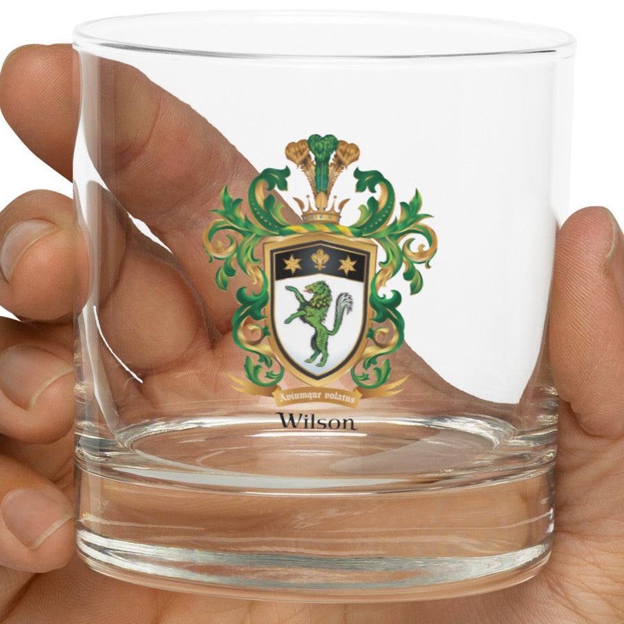Hand holding a clear rocks glass with a Wilson Family Crest green wolf coat of arms design on a white background
