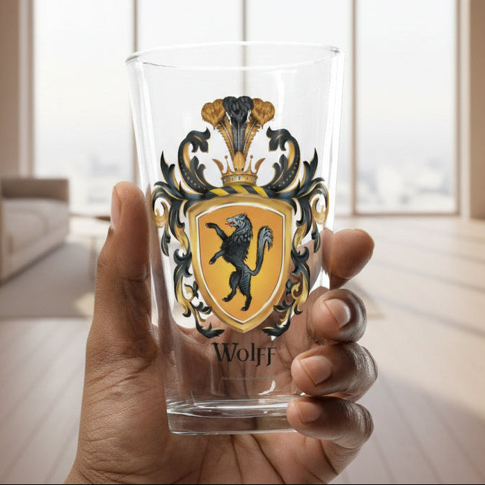 A hand holds a clear drinking glass featuring a printed heraldic crest with a wolf on a golden shield and the name 'Wolff' below, against a softly blurred indoor background.