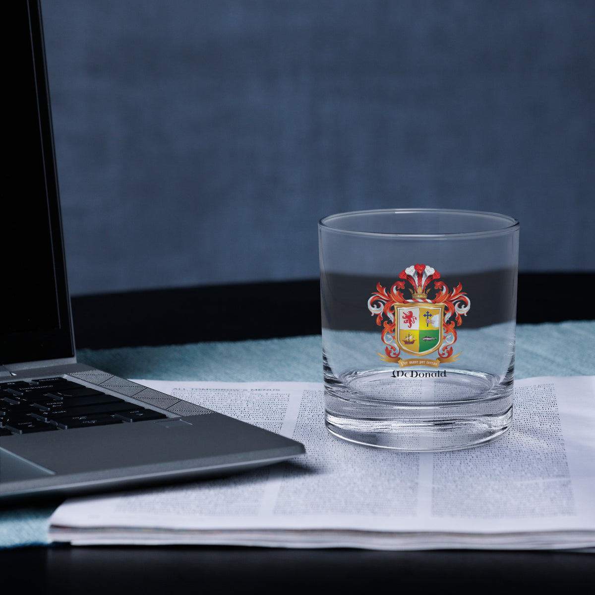Clear glass with a colorful McDonald Family Crest rocks whiskey glass  on a desk next to a laptop