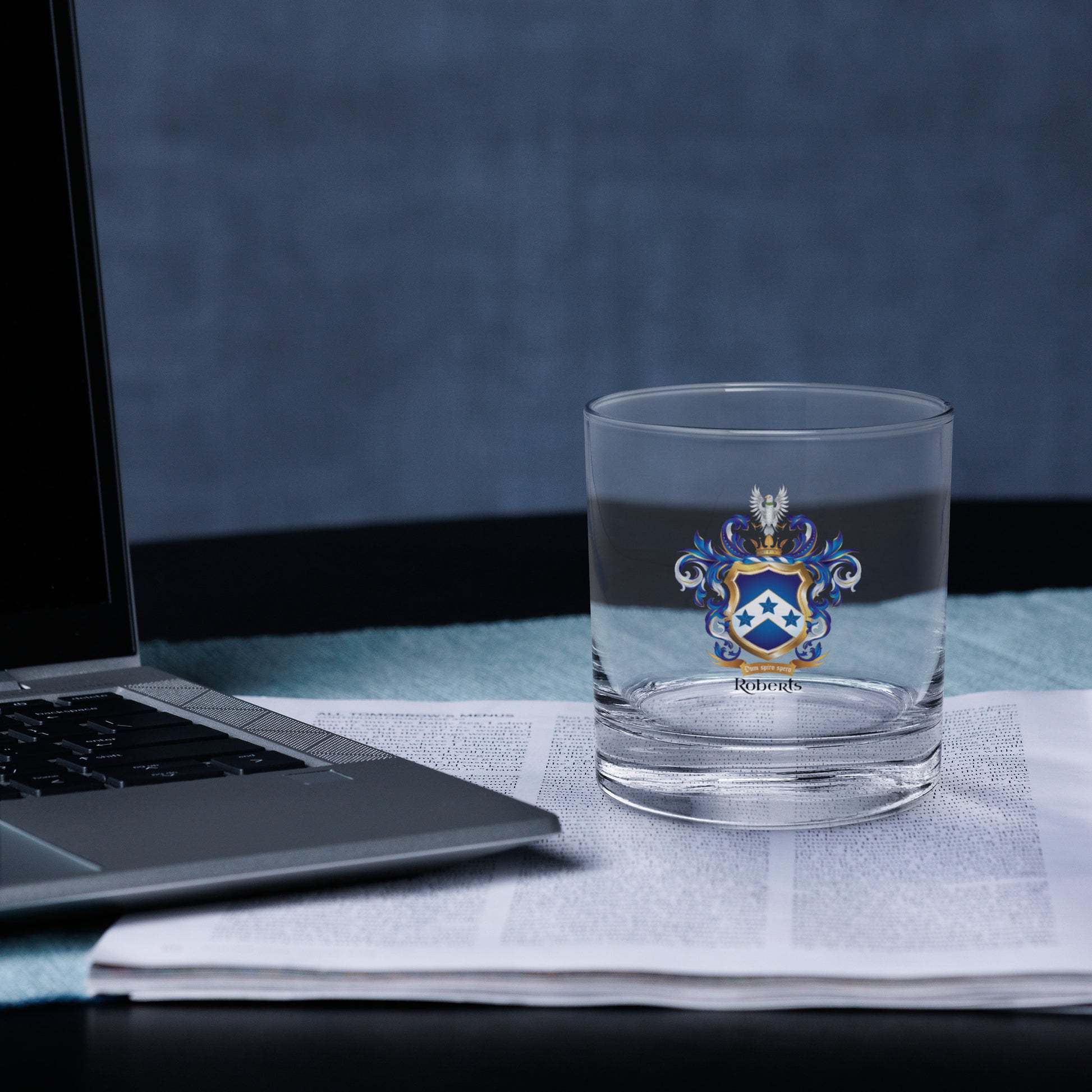 Clear glass with a Roberts  coat of arms design on a desk next to a laptop.
